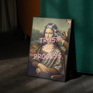 May include: Framed art print depicting a parody of the Mona Lisa having her hair styled. The image includes the pink text "TRUST THE PROCESS". The artwork is set against a landscape backdrop and is framed in a black border.