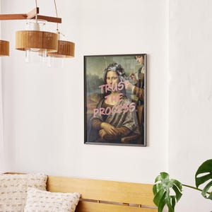 May include: A framed print of the Mona Lisa with the words "Trust the Process" in pink. The artwork hangs in a room with a wooden bench, cushions, and a modern light fixture with woven shades. The room has a minimalist design.