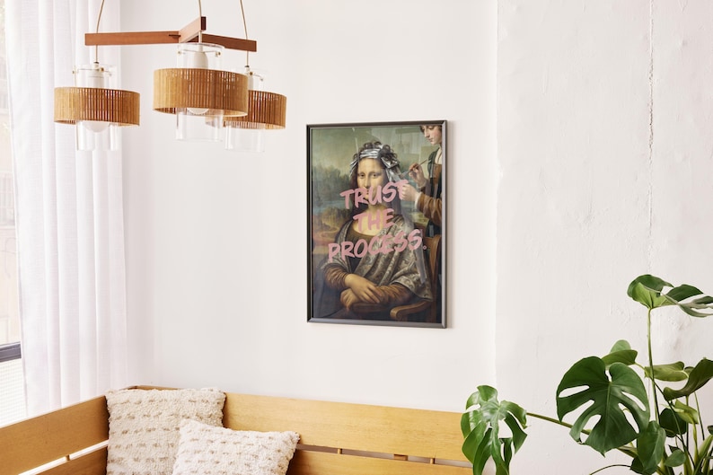 May include: A framed print of the Mona Lisa with the words "Trust the Process" in pink. The artwork hangs in a room with a wooden bench, cushions, and a modern light fixture with woven shades. The room has a minimalist design.