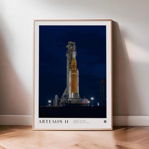 Op de afbeelding: Een ingelijste print met de Artemis II-raket op de lanceerbasis 's nachts. De raket is oranje en wit, met de Amerikaanse vlag zichtbaar. De print bevat de tekst "ARTEMIS II" onderaan.