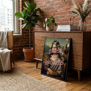 May include: Framed art print showing a parody of the Mona Lisa with the text "Trust the Process" in pink. The artwork depicts the Mona Lisa with hair foils, receiving a beauty treatment. The print is displayed on a wooden floor.