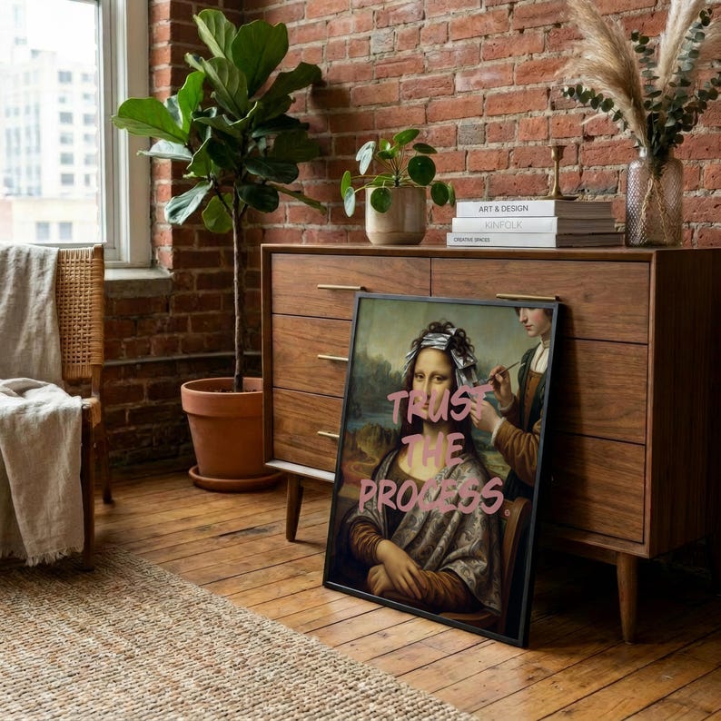 May include: Framed art print showing a parody of the Mona Lisa with the text "Trust the Process" in pink. The artwork depicts the Mona Lisa with hair foils, receiving a beauty treatment. The print is displayed on a wooden floor.
