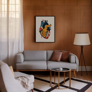May include: A framed anatomical heart print in a living room setting. The artwork features a colorful illustration of a human heart with red, yellow, blue, and black hues. The room includes a gray sofa, a side table, and a floor lamp.