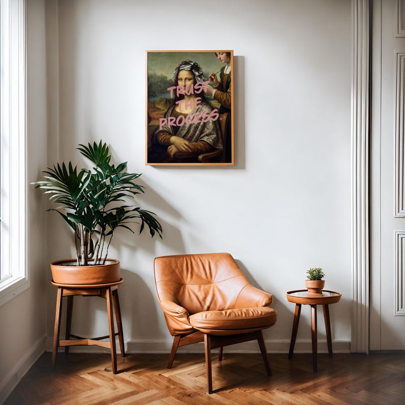 May include: A framed artwork of the Mona Lisa with the text "Trust the Process" in pink. The artwork hangs on a white wall above a brown leather armchair and a small side table. A potted plant sits on a wooden stand.