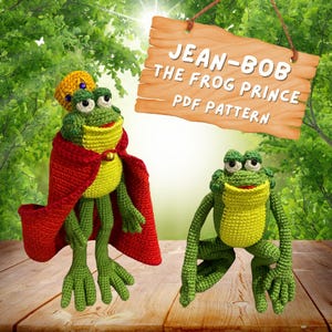 May include: Two crocheted frog plushies, one wearing a red cape and a gold crown, the other sitting on a wooden surface. The sign above reads "Jean-Bob The Frog Prince PDF Pattern".