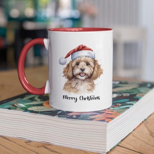 May include: White ceramic mug with a red handle and red rim. The mug features a watercolor illustration of a brown and white dog wearing a red Santa hat. The text "Merry Christmas" is printed in black below the dog.