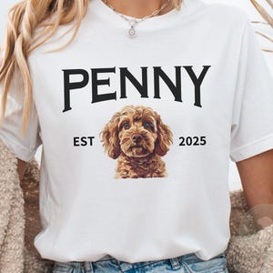 Op de afbeelding: Wit T-shirt met de naam "PENNY" in zwart, boven een gedetailleerde illustratie van een bruine hond. Onder de hond staat de tekst "EST 2025". Het T-shirt is een casual stijl.