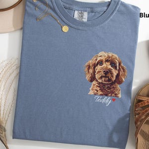 Puede incluir: Una camiseta azul vaquero con una ilustración de un perro marrón y blanco y el texto "Teddy" en la parte delantera.