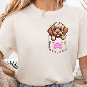 Puede incluir: Camiseta color crema con un perro de dibujos animados asomando por un bolsillo. El perro es marrón y canela con una etiqueta rosa en forma de hueso que dice "DAISY". El bolsillo está delineado en negro.
