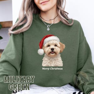 May include: Military green sweatshirt with a dog wearing a Santa hat. The dog illustration has a white and tan coat and is smiling. The text "Merry Christmas" is below the dog. The words "MILITARY GREEN" are printed at the bottom.