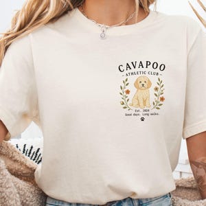 May include: A cream-coloured t-shirt with a graphic of a Cavapoo dog inside a floral wreath. The text reads "Cavapoo Athletic Club" with "Est. 2024" and "Good days. Long walks." printed below. A paw print is also included.