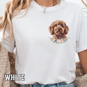 Op de afbeelding: Wit T-shirt met een aquarel illustratie van een bruine Cockapoo hond met een bloemenkraag. De hond is omringd door een krans van pastelkleurige bloemen. Het woord "WHITE" staat onderaan het shirt.