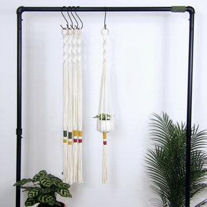 Op de afbeelding: Vijf witte macramé plantenhangers met gele, rode en groene accenten die aan een zwarte metalen rek hangen. Een hanger houdt een kleine plant in een pot.
