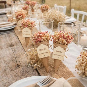 May include: A wedding table decorated with cone-shaped floral arrangements. Each cone contains dried flowers in pink and beige, with a wooden tag saying "Thank You". The table is set with plates, glasses, and cutlery, creating a rustic aesthetic.