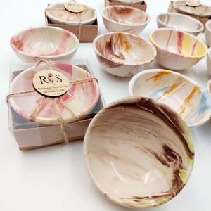 Puede incluir: Una colección de pequeños cuencos de cerámica decorativos con diseños en espiral en varios colores, incluyendo rosa, marrón, amarillo y azul. Algunos cuencos están empaquetados con etiquetas de madera que dicen "Rosemary and Samuel October 21, 2023".