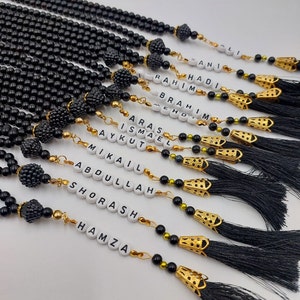 May include: A collection of black prayer beads with gold accents and tassels. Each bead strand has a white tag with a different name on it, including "Hadi", "Rahim", "Ibrahim", "Aras", "Ismael", "Aykut", "Mikail", "Abdullah", "Shorash", and "Hamza".