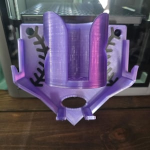 May include: A 3D-printed, purple and lavender-colored object, possibly a holder or mount. The design features a central cylindrical section flanked by curved supports and decorative cutouts. The object is set against a blurred background, suggesting a workshop or manufacturing environment.