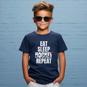 May include: A navy blue t-shirt with white text that reads "Eat Sleep Hockey Repeat". The shirt features a graphic of hockey players.