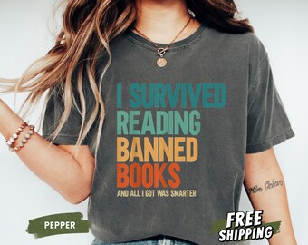 Puede incluir: Camiseta gris oscuro con el texto "I SURVIVED READING BANNED BOOKS AND ALL I GOT WAS SMARTER" en verde azulado, naranja y blanco. La camiseta también tiene la palabra "PEPPER" y "FREE SHIPPING" en verde.