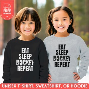 May include: Two children wearing sweatshirts with the text "Eat Sleep Hockey Repeat" printed on them. The sweatshirts are black and gray. The children are smiling.