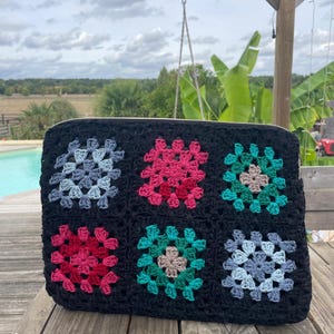 Peut inclure: Sac en crochet noir avec six carrés fleuris colorés. Les carrés présentent des nuances de rose, de bleu et de vert. Le sac est posé sur une surface en bois, avec une piscine et de la verdure en arrière-plan.