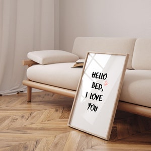 Puede incluir: Una impresión enmarcada con el texto "Hello Bed, I Love You" en letras negras con un corazón rosa. La impresión se apoya en un sofá beige claro con patas de madera.