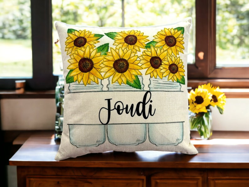 15 x 15 pillow with white background with a printed image of three mason jars filled with yellow sunflowers with a custom family name in a cutout in the mason jars in a script font and black ink