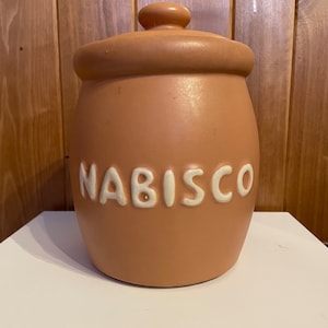 Pode incluir: Um pote de biscoitos de cerâmica cor terracota com uma tampa branca e a palavra "NABISCO" impressa em letras brancas na frente.