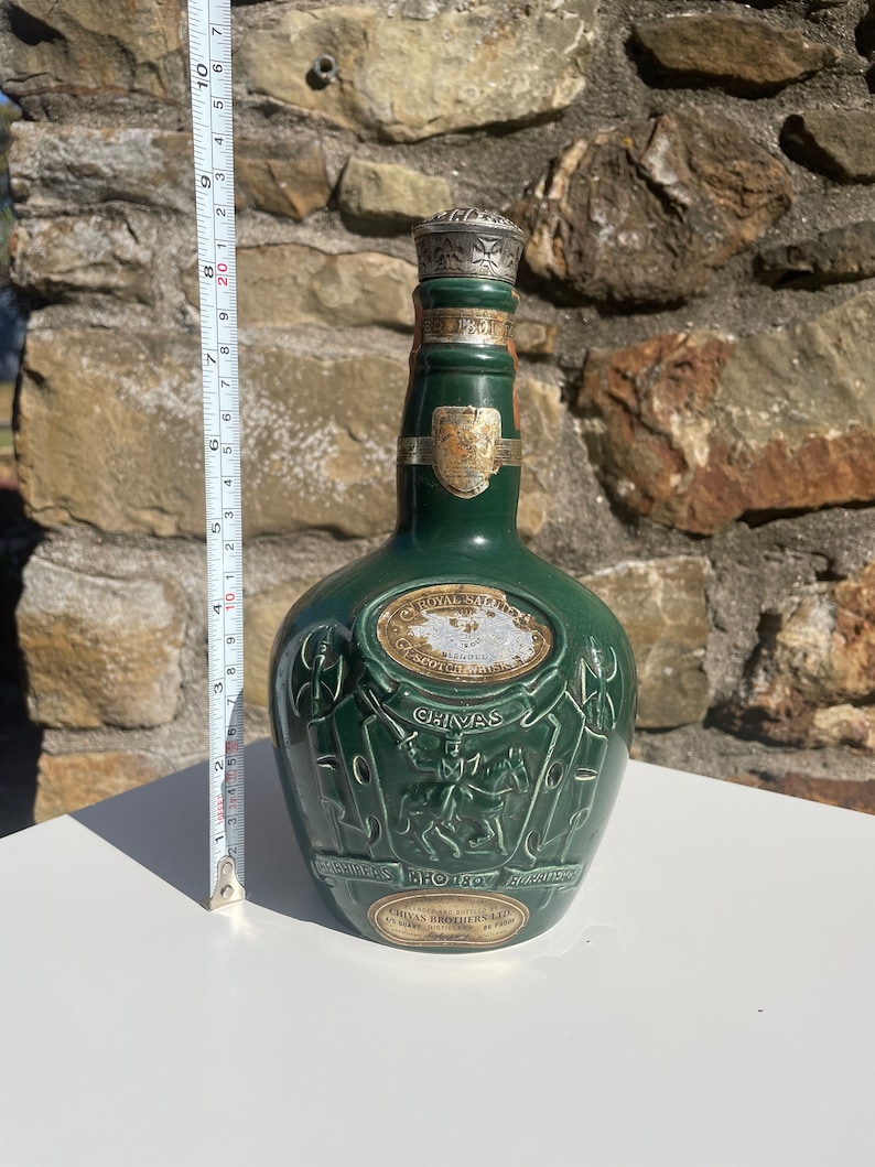 May include: A green glass bottle of Chivas Regal Scotch Whisky with a silver top. The bottle has a label with the words "Chivas Regal" and "Scotch Whisky" on it. The bottle is sitting on a white surface.
