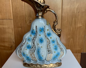 May include: A white ceramic decanter with a gold-colored spout and handle. The decanter is decorated with blue and gold floral designs.
