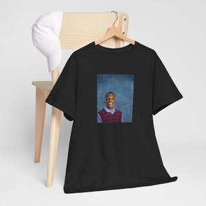 Op de afbeelding: Zwart T-shirt met een foto van een persoon die een rood-witte geruite trui draagt. De persoon glimlacht en heeft kort zwart haar.