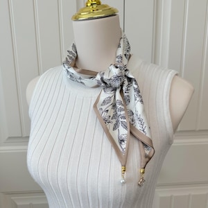 May include: A white and beige silk scarf with a floral pattern and a brown border is tied around a mannequin's neck. The scarf has a knot in the center and is adorned with pearl and crystal charms.