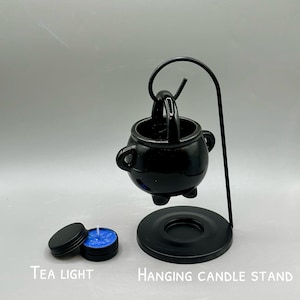 May include: Black hanging candle stand with a cauldron-shaped candle holder. Includes a small, round tea light with a blue candle and a black lid. The words "Tea Light" and "Hanging Candle Stand" are printed on the image.