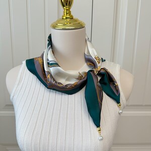 May include: A silk scarf with a white background and a pattern of gold, green, and gray designs. The scarf is draped around a mannequin's neck and has green tips with pearl accents.