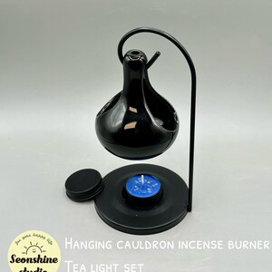 May include: Black hanging cauldron incense burner with a tea light set. The burner is suspended by a curved metal stand. The set includes a black base, a lid, and a blue tea light candle. Text on the image reads "Hanging Cauldron Incense Burner Tea Light Set."