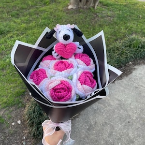 May include: A handmade crochet bouquet featuring pink and white roses, a Snoopy dog with a pink heart, and black and white wrapping. The bouquet is tied with a sheer white ribbon.
