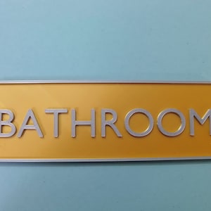 May include: A yellow rectangular sign with a silver border and the word "BATHROOM" in silver lettering.
