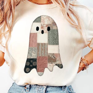 May include: Cream-colored t-shirt featuring a patchwork ghost design. The ghost is composed of various fabric patterns in shades of pink, gray, and black. The shirt has a relaxed fit and short sleeves, suitable for casual wear.