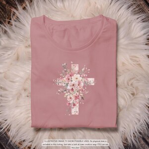 以下が含まれることがあります： ピンクがかったバラ色のTシャツ。花の十字架のデザインが特徴です。十字架はピンクと白の花と緑の葉で飾られています。シャツは折りたたまれ、ふわふわの白い表面に置かれています。下部のテキストは次のとおりです：ILLUSTRATIVE IMAGE TO SHOW POSSIBLE USES.
