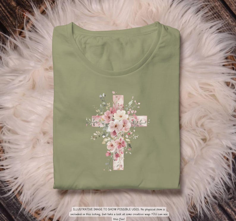 以下が含まれることがあります： セージグリーンのTシャツ。花の十字架のデザインが特徴です。十字架はピンクと白の花と緑の葉で飾られています。Tシャツはふわふわの白い表面に折りたたまれています。下部のテキストは次のとおりです: ILLUSTRATIVE IMAGE TO SHOW POSSIBLE USES.