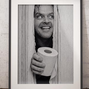 Pode incluir: Uma impressão emoldurada a preto e branco mostrando um homem com uma expressão intensa a espreitar por uma fenda vertical numa porta de madeira, segurando um rolo de papel higiénico. A impressão está numa moldura preta simples.