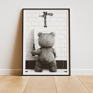May include: Framed monochrome print depicting a teddy bear in front of a urinal, holding a bottle. The background shows a tiled wall and a chrome fixture. The print is displayed on a wooden floor.
