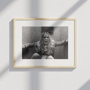 Puede incluir: Impresi&oacute;n enmarcada en blanco y negro que presenta a una persona con cabello largo y rubio, sentada y con aspecto angustiado. La imagen es un primer plano, con los brazos de la persona extendidos. La impresi&oacute;n est&aacute; en un marco de madera de color claro.