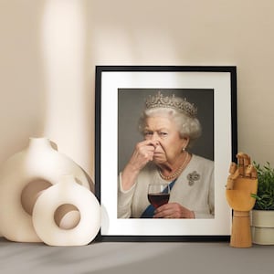 May include: Framed print featuring a portrait of Queen Elizabeth II, holding a glass of red wine and pinching her nose. The image is in a black frame, with a white mat. Decorative vases and a wooden hand sculpture are in the foreground.