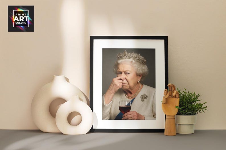 May include: Framed print featuring a portrait of Queen Elizabeth II, holding a glass of red wine and pinching her nose. The image is in a black frame, with a white mat. Decorative vases and a wooden hand sculpture are in the foreground.