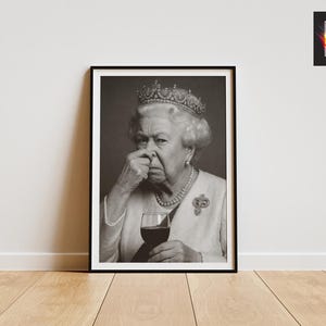 May include: Black and white framed print featuring a portrait of Queen Elizabeth II. The image shows the Queen holding a glass of red wine and pinching her nose. The print is displayed on a wooden floor against a white wall.