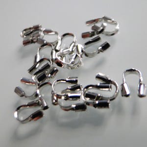 May include: A collection of silver-coloured crimp end findings, designed for jewellery making. These small, U-shaped metal pieces have a tube-like end for securing cords or ribbons. The findings are scattered on a light grey surface.