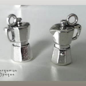May include: Two small, silver-colored coffee maker charms. Each charm has a handle, a spout, and a loop at the top for attaching to a chain or bracelet. The charms are miniature replicas of a stovetop espresso maker.