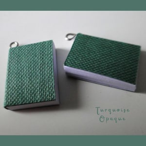 Könnte beinhalten: Zwei Miniatur-Buchanhänger in Türkisgrün mit strukturierten Einbänden und silberfarbenen Schlaufen. Die Bücher haben weiße Seiten und sind vor einem hellen Hintergrund platziert. Der Text "Turquoise Opaque" ist sichtbar.