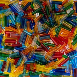 May include: A collection of translucent, cylindrical glass tube beads in a vibrant assortment of colours including red, blue, green, yellow, and orange. Each bead features a hollow centre and a smooth, glossy finish, scattered in a dense pile.